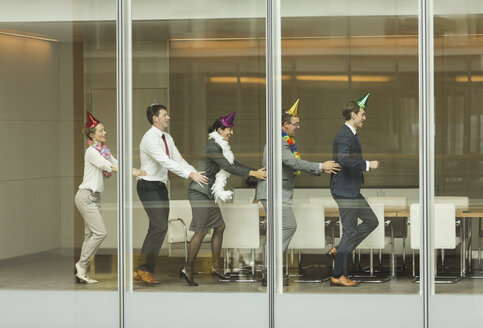 Geschäftsleute mit Partyhüten tanzen in einer Conga-Linie am Fenster des Konferenzraums - CAIF06705