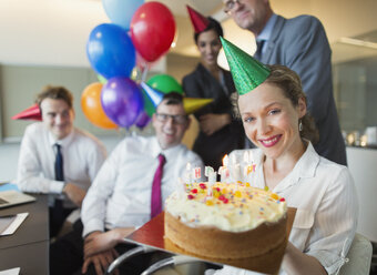 Porträt lächelnde Geschäftsfrau hält Geburtstagstorte mit Kollegen im Hintergrund - CAIF06709