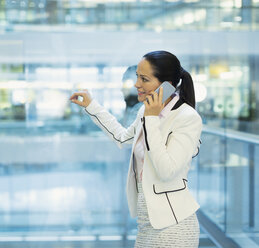 Businesswoman talking on cell phone at office window - CAIF06740