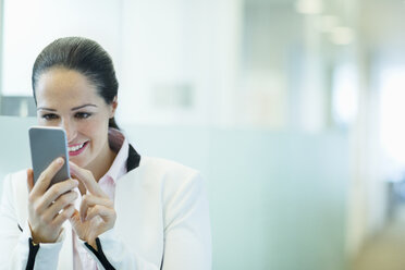 Lächelnde Geschäftsfrau, die im Büroflur ein Mobiltelefon benutzt - CAIF06766