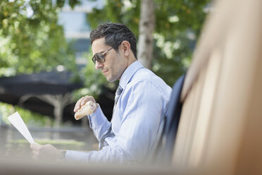 Corporate businessman eating lunch and reading paperwork on park bench - CAIF06821