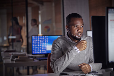 Businessman working late at computer, using hands-free headphones talking on telephone - CAIF06876