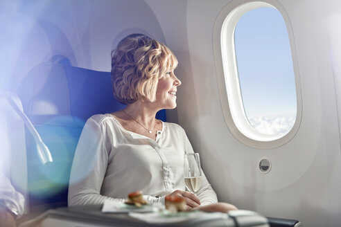 Lächelnde Frau, die Champagner trinkt, in der ersten Klasse reist und aus dem Flugzeugfenster schaut - CAIF06983