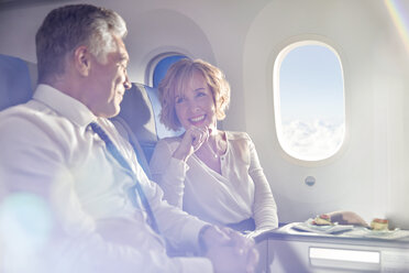 Lächelndes reifes Paar, das in der ersten Klasse eines Flugzeugs isst und spricht - CAIF06991