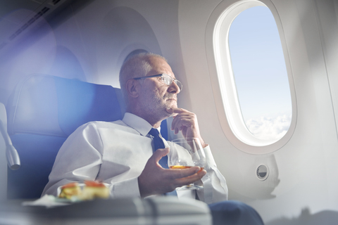 Älterer Geschäftsmann trinkt Whiskey in der ersten Klasse und schaut aus dem Flugzeugfenster, lizenzfreies Stockfoto