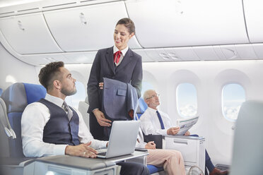 Flight attendant talking with businessman working at laptop on airplane - CAIF07043