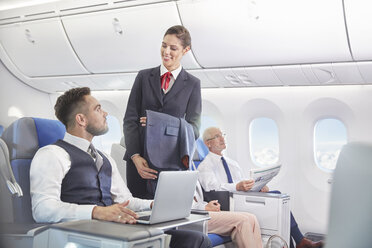Flugbegleiter im Gespräch mit einem Geschäftsmann, der im Flugzeug an einem Laptop arbeitet - CAIF07043