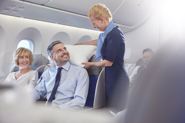 Lächelnde Flugbegleiterin passt Kissen für Geschäftsmann in der ersten Klasse im Flugzeug an - CAIF07049