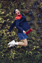 Porträt einer modischen jungen Frau, die vor der Living Wall in die Luft springt - JSMF00085