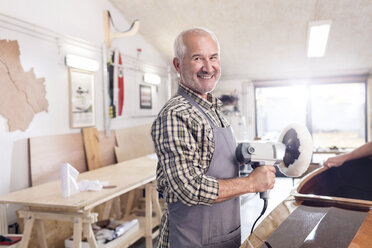 Portrait smiling, confident senior male carpenter using a buffer sander on wood boat in workshop - CAIF07059