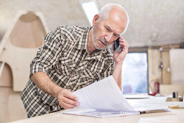 Senior carpenter talking on cell phone and reviewing paperwork in workshop - CAIF07064