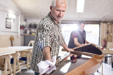 Senior man finishing, wiping wood kayak in workshop - CAIF07066