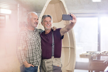 Stolze, lächelnde männliche Tischler mit Fotohandy machen ein Selfie neben einem Holzboot in der Werkstatt - CAIF07072