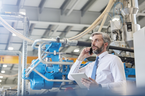 Männlicher Vorgesetzter mit digitalem Tablet, der in einer Fabrik mit einem Mobiltelefon spricht, lizenzfreies Stockfoto