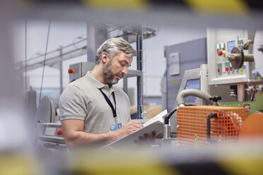 Männlicher Vorgesetzter mit Klemmbrett an einer Maschinenkontrolltafel in einer Fabrik für Glasfasertechnik - CAIF07197