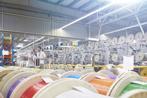 Männlicher Vorgesetzter und Arbeiter überprüfen das Inventar hinter mehrfarbigen Spulen in einer Glasfaserfabrik, lizenzfreies Stockfoto