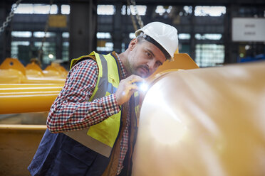 Fokussierter männlicher Ingenieur mit Taschenlampe, der die Ausrüstung in einer Fabrik untersucht - CAIF07256