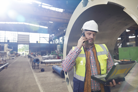 Männlicher Ingenieur mit Laptop, der in einem Stahlwerk mit seinem Handy telefoniert, lizenzfreies Stockfoto