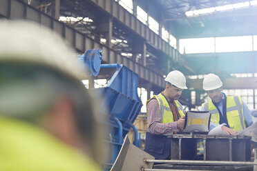 Male foreman and engineer working at laptop, discussing blueprints in factory - CAIF07267