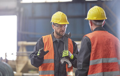 Männlicher Arbeiter mit großem Schraubenschlüssel im Gespräch mit einem Arbeitskollegen in einer Fabrik - CAIF07269