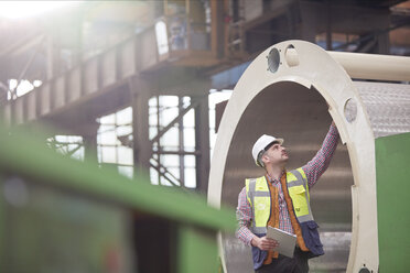 Male engineer with clipboard examining steel pipe in factory - CAIF07271