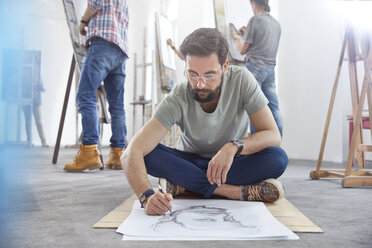 Männlicher Künstler sitzt im Schneidersitz und skizziert auf dem Boden im Atelier eines Kunstkurses - CAIF07294