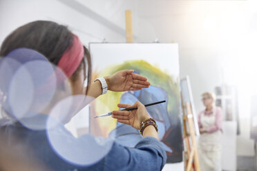 Female artist gesturing, framing painting on easel in art class studio - CAIF07297