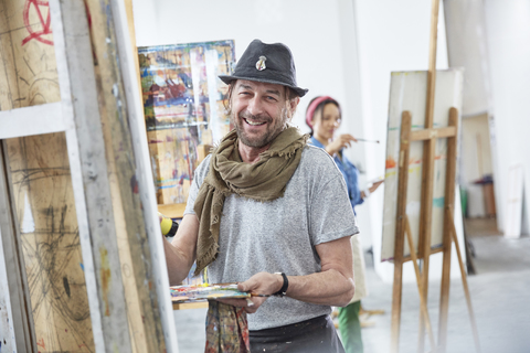 Porträt eines lächelnden männlichen Künstlers, der an einer Staffelei im Atelier eines Kunstkurses malt, lizenzfreies Stockfoto