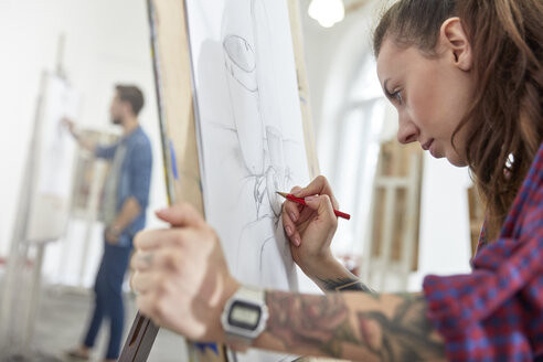 Fokussierte Künstlerin mit Tätowierung, die an einer Staffelei im Atelier des Kunstunterrichts skizziert - CAIF07321