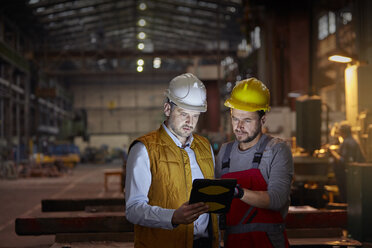 Male foreman and engineer using digital tablet in dark factory - CAIF07337