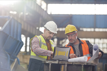 Male foreman and engineer working at laptop with blueprints in factory - CAIF07340
