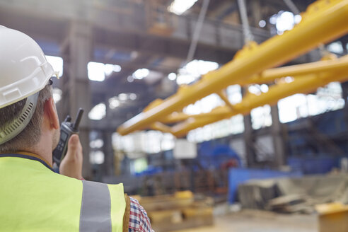 Männlicher Arbeiter benutzt ein Walkie-Talkie, um eine hydraulische Kranabsenkvorrichtung in einer Fabrik zu steuern - CAIF07362