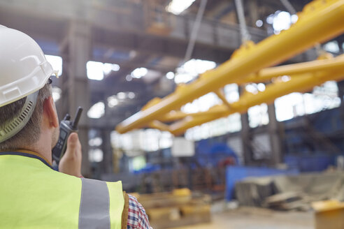 Male worker using walkie-talkie to guide hydraulic crane lowering equipment in factory - CAIF07362