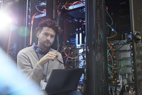 Porträt eines selbstbewussten männlichen IT-Technikers, der in einem Serverraum an einem Laptop arbeitet - CAIF07397