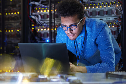 Fokussierter männlicher IT-Techniker, der in einem dunklen Serverraum an einem Laptop arbeitet - CAIF07416
