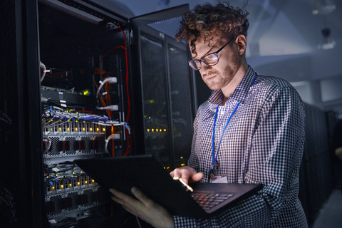 Fokussierter männlicher IT-Techniker, der in einem dunklen Serverraum an einem Laptop arbeitet, lizenzfreies Stockfoto