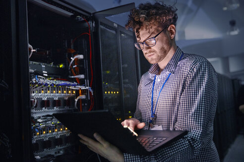 Fokussierter männlicher IT-Techniker, der in einem dunklen Serverraum an einem Laptop arbeitet - CAIF07424