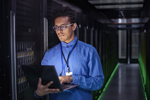 Fokussierter männlicher IT-Techniker mit Laptop in einem dunklen Serverraum - CAIF07431