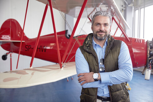 Porträt eines lächelnden, selbstbewussten männlichen Flugzeugpiloten, der an einem Doppeldecker in einem Hangar steht - CAIF07456