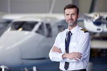 Porträt eines selbstbewussten männlichen Piloten, der in der Nähe eines Flugzeugs in einem Hangar steht - CAIF07462