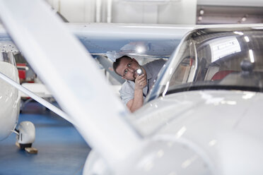 Ein Mechaniker untersucht die Unterseite eines Flugzeugflügels mit einer Taschenlampe im Hangar - CAIF07478