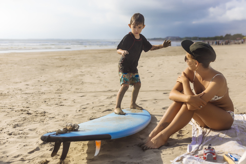 Indonesien, Bali, Junge steht auf Surfbrett, Mutter sitzt am Strand, lizenzfreies Stockfoto