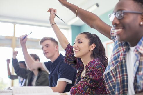 Schüler sitzen im Klassenzimmer während des Unterrichts, lächeln und heben die Hände - CAIF07695