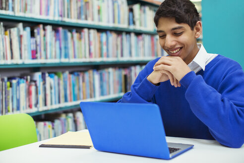 Porträt eines lächelnden Studenten, der in der Bibliothek auf einen Laptop schaut - CAIF07707