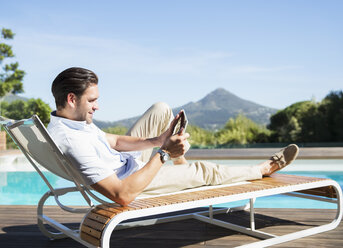 Mann mit digitalem Tablet auf einem Liegestuhl am Pool - CAIF07858