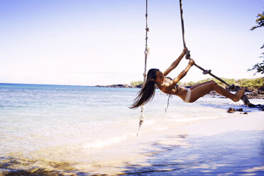 Woman in bikini swinging on rope at beach against clear sky - CAVF01444