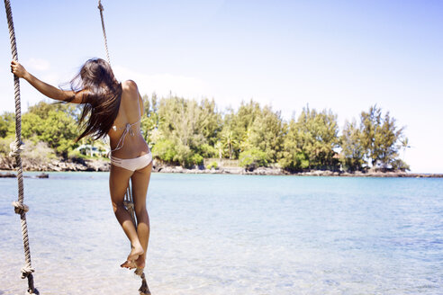 Rückansicht einer Frau im Bikini, die an einem Seil am Strand schwingt - CAVF01445