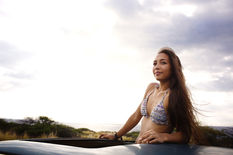 Frau schaut weg, während sie im Schiebedach eines Autos gegen den Himmel steht, lizenzfreies Stockfoto