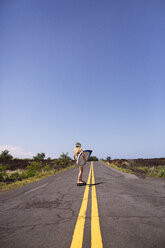 Woman carrying surfboard while skateboarding on road against blue sky - CAVF01484