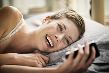 Smiling woman looking at smart phone while lying on bed - CAVF01518